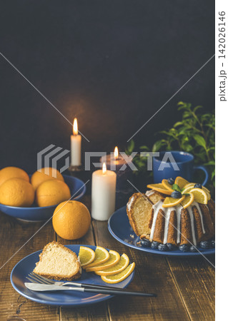 Orange bundt cake with blueberry surrounded fruits, candles, plant and cutleries on dark wooden background. Orange bundt cake with blueberry surrounded fruits, candles, plant and cutleries on dark wooden background. 142026146