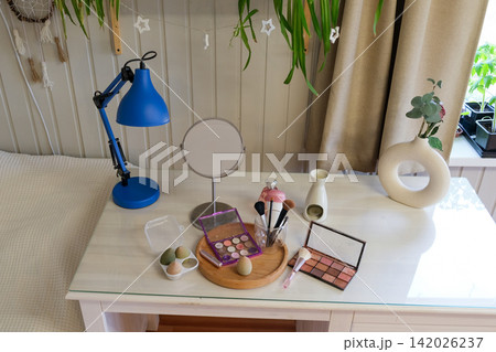 Vanity round mirror and make up items arranged on desk under lamp, palettes and brushes, open eyeshadow, cotton pads. Makeup and self care at home concept. Personal beauty products items on table. 142026237