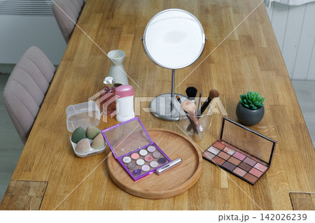 Vanity round mirror and make up items arranged on desk under lamp, palettes and brushes, open eyeshadow, cotton pads. Makeup and self care at home concept. Personal beauty products items on table. 142026239