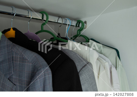 Jackets, black coats, and light-colored blouses hang side by side on plastic and wire hangers against a white wall. Concept: wardrobe density, contrast of textures and colors. High quality photo Jackets, black coats, and light-colored blouses hang side by side on plastic and wire hangers against a white wall. Concept: wardrobe density, contrast of textures and colors. High quality photo 142027808