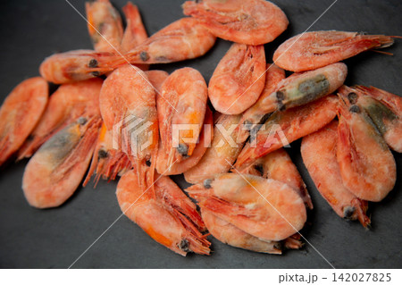 A close-up of frozen shrimps covered with white frost are scattered on a black stone surface, their pink-orange shell and dark eyes stand out. Concept: detailing the texture of frozen. High quality 142027825