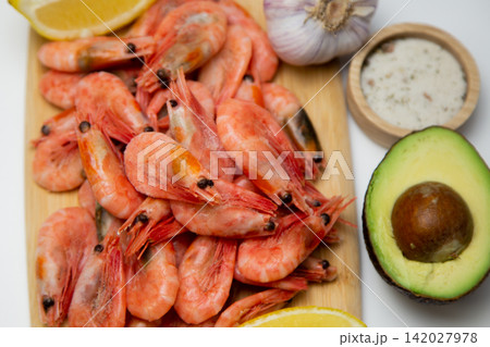 A close-up of a wooden board with frozen shrimps, lemon slices, half an avocado, garlic and a salt shaker are located next to it. Concept: detailing the texture and color of seafood. High quality A close-up of a wooden board with frozen shrimps, lemon slices, half an avocado, garlic and a salt shaker are located next to it. Concept: detailing the texture and color of seafood. High quality 142027978