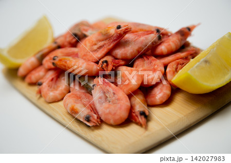 Close-up of frozen pink shrimp on a light wooden board surrounded by two bright lemon slices on a white background. Concept: bright color contrast, gastronomic aesthetics of fresh seafood. High 142027983
