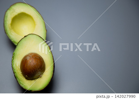 On a gray background, two avocado halves are located on the left side of the frame, the lower one with a stone, the upper one without it, creating a vertical composition. Concept: minimalistic 142027990