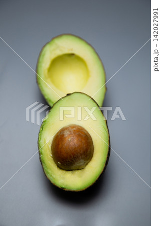 Vertical view of two avocado halves: the lower one with a large round stone, the upper one with an empty notch on a smooth gray background. Concept: aesthetics of simple composition, structure and 142027991