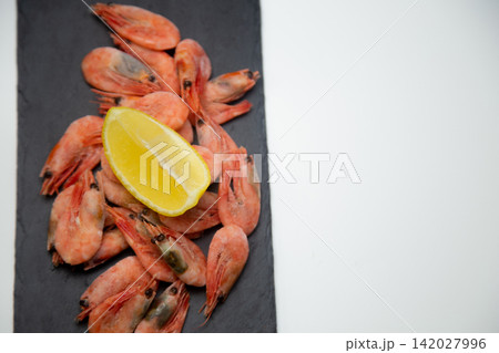 A close-up of a handful of frozen shrimp on a black stone board with textured uneven edges, on top of them lies a slice of lemon with space for text. Concept: micro-detailing of seafood. High quality A close-up of a handful of frozen shrimp on a black stone board with textured uneven edges, on top of them lies a slice of lemon with space for text. Concept: micro-detailing of seafood. High quality 142027996