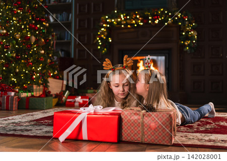 Merry Christmas Xmas holiday. Children unpacking gift box near Christmas tree. Two sister kids in living room with Christmas tree fireplace opening gift box with surprise faces. Christmas eve at home Merry Christmas Xmas holiday. Children unpacking gift box near Christmas tree. Two sister kids in living room with Christmas tree fireplace opening gift box with surprise faces. Christmas eve at home 142028001