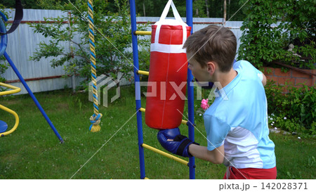 Authentic portrait of a young male athlete boxer hitting a red hanging bag in a residential courtyard area under school holiday health promotion guidelines Authentic portrait of a young male athlete boxer hitting a red hanging bag in a residential courtyard area under school holiday health promotion guidelines 142028371