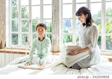 ベッドの上で洗濯物をたたむ若い母親と小学生の女の子 ベッドの上で洗濯物をたたむ若い母親と小学生の女の子 142034609