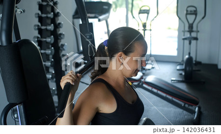 Young woman in black sportswear performing a behind-the-neck lat pulldown exercise on a cable machine in a gym. Back muscle training concept 142036431