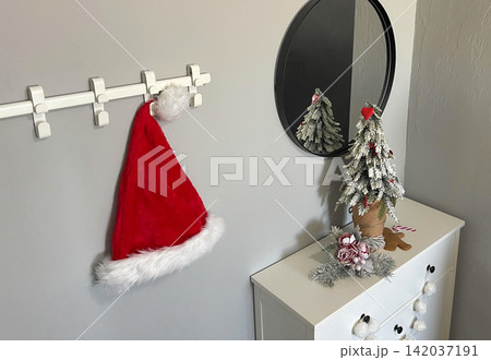 Red Christmas hat hangs on a hook near a round mirror with a small decorative tree Red Christmas hat hangs on a hook near a round mirror with a small decorative tree 142037191