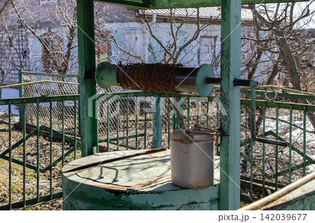 A rustic garden water well with a weathered metal bucket and rusty chain stands in a rural backyard, evoking a nostalgic sense of simple country living and traditional heritage life 142039677
