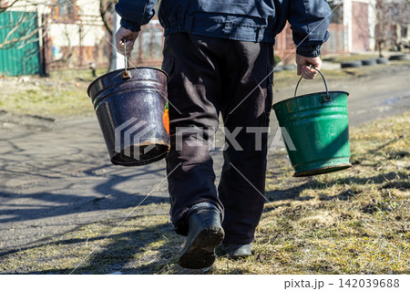 A worker carries two metal buckets while walking along a path, representing labor, daily maintenance, domestic chores, and a manual task within a rural or industrial setting today 142039688