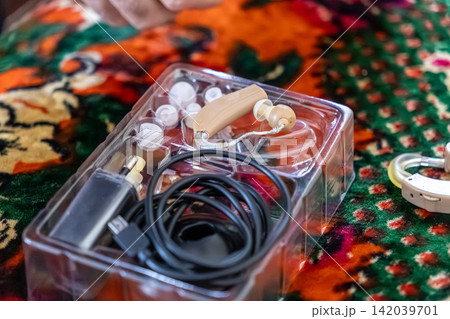 A beige rechargeable hearing aid device lies inside a plastic storage tray with a charging cable and silicone tips to improve audio clarity for individuals with sensory loss issues A beige rechargeable hearing aid device lies inside a plastic storage tray with a charging cable and silicone tips to improve audio clarity for individuals with sensory loss issues 142039701