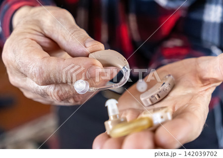 A senior person holds multiple modern hearing aid devices in hands, demonstrating medical equipment for auditory support and health care solutions to improve quality of life for elderly A senior person holds multiple modern hearing aid devices in hands, demonstrating medical equipment for auditory support and health care solutions to improve quality of life for elderly 142039702