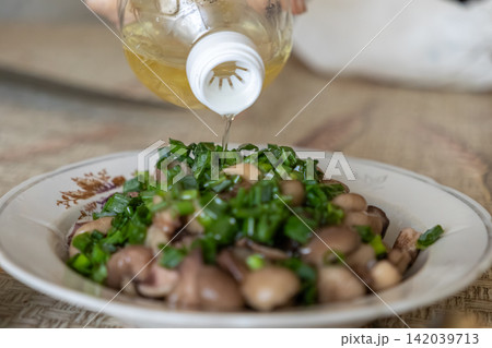 Vegetable oil dripping from a plastic bottle onto a plate of cooked mushrooms with fresh chopped green onions, representing culinary preparation for a healthy homemade meal at home Vegetable oil dripping from a plastic bottle onto a plate of cooked mushrooms with fresh chopped green onions, representing culinary preparation for a healthy homemade meal at home 142039713