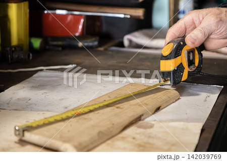 A senior hand uses a yellow metal tape measure to check a pine plank, highlighting precision and measurement in a professional carpentry workshop for home improvement projects 142039769