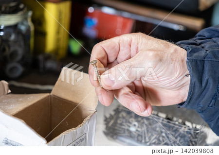 A mature person holds a metal screw between fingers in a cluttered workshop, depicting preparation for home improvement tasks or manual labor projects in a casual residential setting A mature person holds a metal screw between fingers in a cluttered workshop, depicting preparation for home improvement tasks or manual labor projects in a casual residential setting 142039808