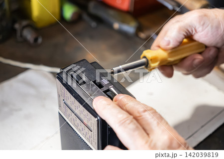 A person uses a yellow screwdriver to repair a black portable radio on a wooden workbench, symbolizing vintage electronics maintenance and technical skills for classic audio repair A person uses a yellow screwdriver to repair a black portable radio on a wooden workbench, symbolizing vintage electronics maintenance and technical skills for classic audio repair 142039819