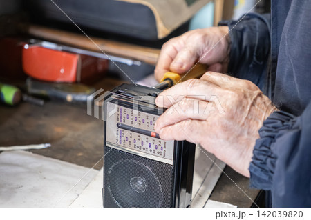 An elderly person repairing a vintage portable radio with a screwdriver in a workshop, conveying technical skill, focused craftsmanship, and the nostalgia of classic electronic repair An elderly person repairing a vintage portable radio with a screwdriver in a workshop, conveying technical skill, focused craftsmanship, and the nostalgia of classic electronic repair 142039820