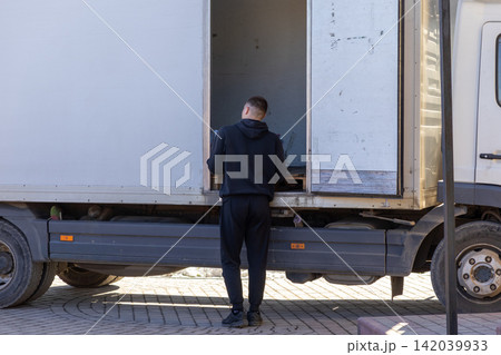 A delivery worker in a black hoodie loads parcels into a white truck, embodying efficiency and logistics services within a professional urban transportation supply chain context A delivery worker in a black hoodie loads parcels into a white truck, embodying efficiency and logistics services within a professional urban transportation supply chain context 142039933