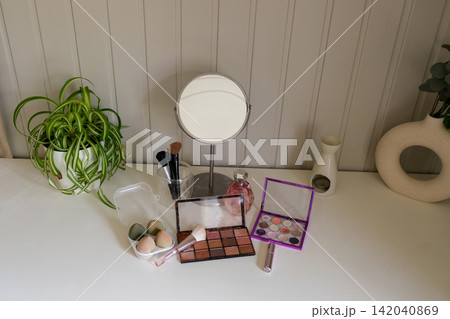 Vanity round mirror and make up items arranged on desk under lamp, palettes and brushes, open eyeshadow, cotton pads. Makeup and self care at home concept. Personal beauty products items on table. 142040869