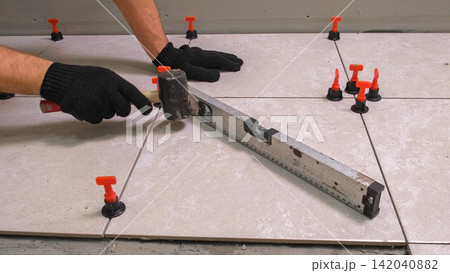 Worker Hand Laying Ceramic Tiles on the Floor. Repairman Placing White Gray Square Tiles, Applying Adhesive. Builder Installing Tile Using Leveler, Tile Leveling Clips. Tiling. Finishing Repair Work. 142040882