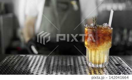 Refreshing layered cocktail with ice and rosemary on bar counter, bartender working in background Refreshing layered cocktail with ice and rosemary on bar counter, bartender working in background 142041664