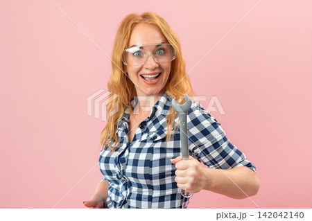 Young woman with red hair wearing a plaid shirt and protective glasses, enthusiastically holding a wrench on a pink background, representing female empowerment in skilled trades 142042140