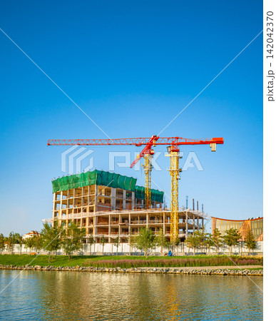 Modern building under construction with crane next to lake, urban development concept Modern building under construction with crane next to lake, urban development concept 142042370