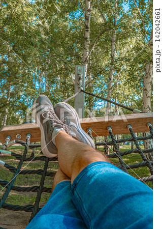 Person resting feet on wooden park net hammock swing in birch tree summer park, outdoor leisure lifestyle concept Person resting feet on wooden park net hammock swing in birch tree summer park, outdoor leisure lifestyle concept 142042661
