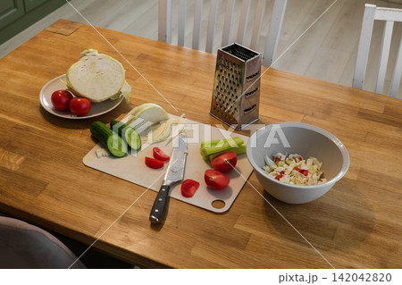 Simple home cooking. Wooden table with fresh vegetables, sliced cabbage, halved tomatoes, cucumber, cutting board knife. Preparing nutritious midday meals. Creating nourishing. Assembling hearty lunch Simple home cooking. Wooden table with fresh vegetables, sliced cabbage, halved tomatoes, cucumber, cutting board knife. Preparing nutritious midday meals. Creating nourishing. Assembling hearty lunch 142042820