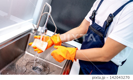 Housekeeper hand wearing orange rubber glove washing sponge under running water from kitchen sink Housekeeper hand wearing orange rubber glove washing sponge under running water from kitchen sink 142043270
