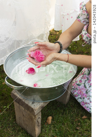 A rustic scene featuring a person in a floral dress holding a handmade soap bar wrapped in white cloth over a metal bucket. 142043928