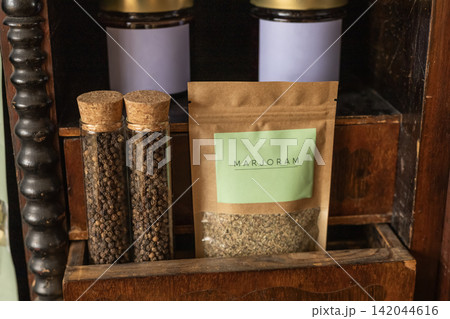 Marjoram spice in kraft pouch and black peppercorns in glass tubes on wood shelf Marjoram spice in kraft pouch and black peppercorns in glass tubes on wood shelf 142044616