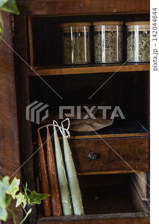 Handmade wax taper candles and dried herbs jars in vintage wooden cabinet 142044644