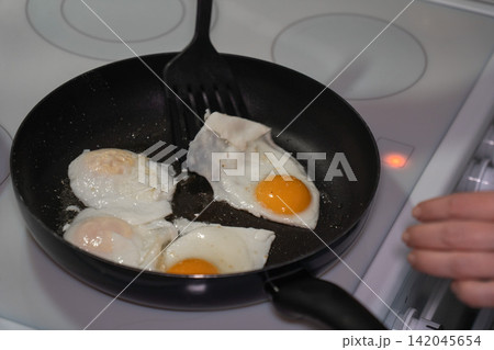 Fried eggs cook in black frying pan while hand uses spatula on stovetop. Home cooking, breakfast preparation, everyday food, kitchen routine, simple meal, domestic lifestyle. Fried eggs cook in black frying pan while hand uses spatula on stovetop. Home cooking, breakfast preparation, everyday food, kitchen routine, simple meal, domestic lifestyle. 142045654