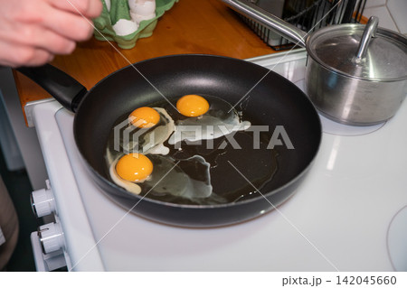 Hand cooks three sunny side up eggs in black frying pan on electric stovetop. Breakfast preparation, home cooking, homemade meal, simple nutrition, kitchen routine, everyday food. Hand cooks three sunny side up eggs in black frying pan on electric stovetop. Breakfast preparation, home cooking, homemade meal, simple nutrition, kitchen routine, everyday food. 142045660
