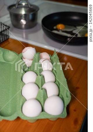 White eggs in open green carton sit beside frying pan on kitchen stovetop. Fresh ingredients, breakfast preparation, home cooking, simple nutrition, homemade meal, everyday kitchen routine. 142045669