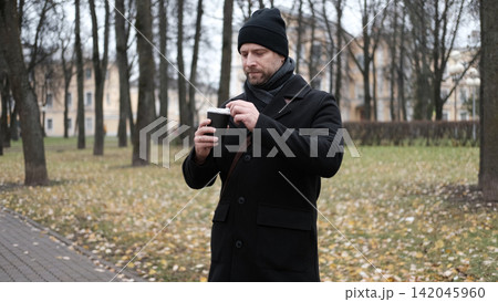 Pensive hipster drinking coffee in the urban street. Handsome man wearing black coat, brown leather bag walking confidently down the city with paper cup in hand. Stylish bearded middle-aged male. 142045960