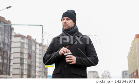 Pensive hipster drinking coffee in the urban street. Handsome man wearing black coat, brown leather bag walking confidently down the city with paper cup in hand. Stylish bearded middle-aged male. 142045971