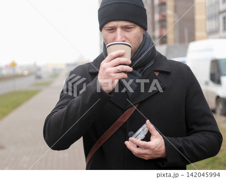 Pensive hipster drinking coffee in the urban street. Handsome man wearing black coat, brown leather bag walking confidently down the city with paper cup in hand. Stylish bearded middle-aged male. Pensive hipster drinking coffee in the urban street. Handsome man wearing black coat, brown leather bag walking confidently down the city with paper cup in hand. Stylish bearded middle-aged male. 142045994