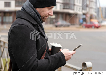 Handsome man wearing black coat and brown leather bag walking down on the city, drinking coffee paper takeaway cup with mobile phone. Stylish bearded middle-aged male using smartphone at urban street Handsome man wearing black coat and brown leather bag walking down on the city, drinking coffee paper takeaway cup with mobile phone. Stylish bearded middle-aged male using smartphone at urban street 142046023