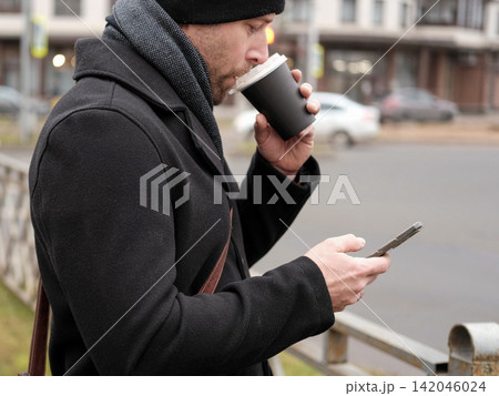 Handsome man wearing black coat and brown leather bag walking down on the city, drinking coffee paper takeaway cup with mobile phone. Stylish bearded middle-aged male using smartphone at urban street Handsome man wearing black coat and brown leather bag walking down on the city, drinking coffee paper takeaway cup with mobile phone. Stylish bearded middle-aged male using smartphone at urban street 142046024