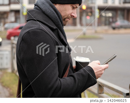 Handsome man wearing black coat and brown leather bag walking down on the city, drinking coffee paper takeaway cup with mobile phone. Stylish bearded middle-aged male using smartphone at urban street 142046025