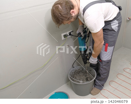 Construction worker preparing tile surface, mixing mortar substrate for flooring, underfloor heating mat. Workman combines adhesive in renovation setting. Operating power tool during bathroom upgrade Construction worker preparing tile surface, mixing mortar substrate for flooring, underfloor heating mat. Workman combines adhesive in renovation setting. Operating power tool during bathroom upgrade 142046079