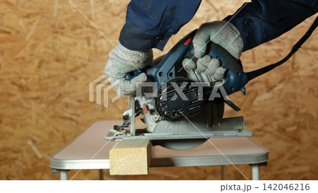 Builder saws a board with a circular saw cutting a wooden plank in a joinery. Carpenter uses portable circular saw tool to cut wood at wooden table in workshop. Worker sawing pine board. Real image 142046216