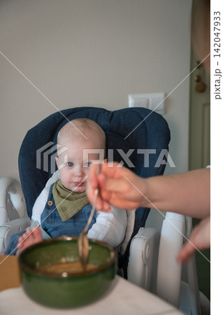 Baby watches spoon as mother offers food while he sits in high chair with bowl at home. Infant feeding, childcare, nutrition, complementary food, curiosity, parenting, development, healthy eating. 142047933