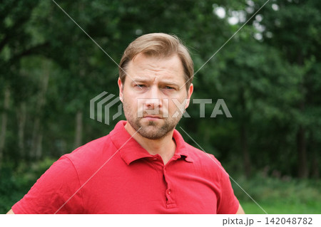 Portrait of handsome young man in a red t-shirt outdoors. Male with fair hair and beard feel happy, calm and serious. Green nature background. Guy looking away into distance. Diversity People Portrait of handsome young man in a red t-shirt outdoors. Male with fair hair and beard feel happy, calm and serious. Green nature background. Guy looking away into distance. Diversity People 142048782