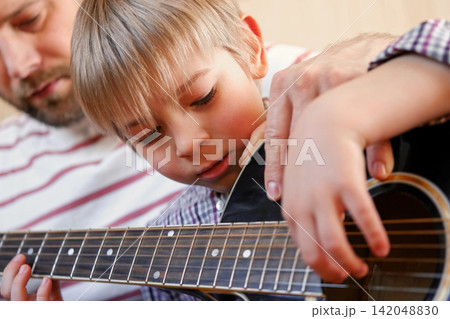Handsome dad teaches his cute son to play the guitar sitting on sofa in the living room at home. Happy Father's Day. Father teaching his child to play guitar. Kid Learning music skill. Art education Handsome dad teaches his cute son to play the guitar sitting on sofa in the living room at home. Happy Father's Day. Father teaching his child to play guitar. Kid Learning music skill. Art education 142048830